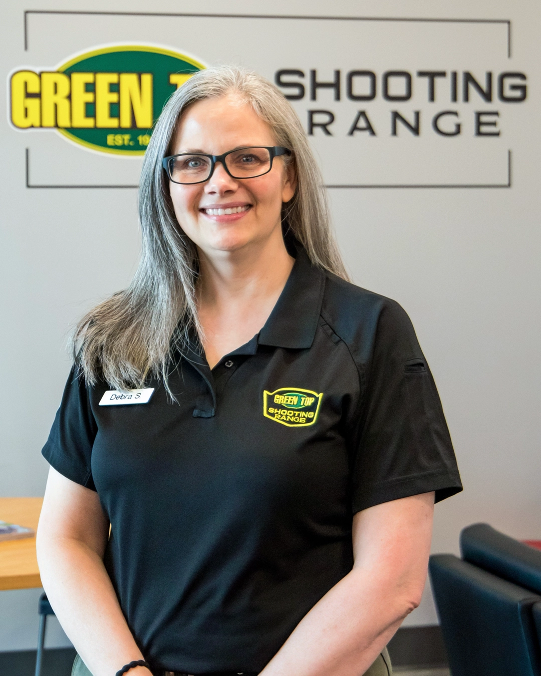 Debra Smith, General Manager at Green Top Shooting Range, with long gray hair and glasses, wearing a black polo with the range logo and smiling in front of the Green Top Shooting Range logo wall.