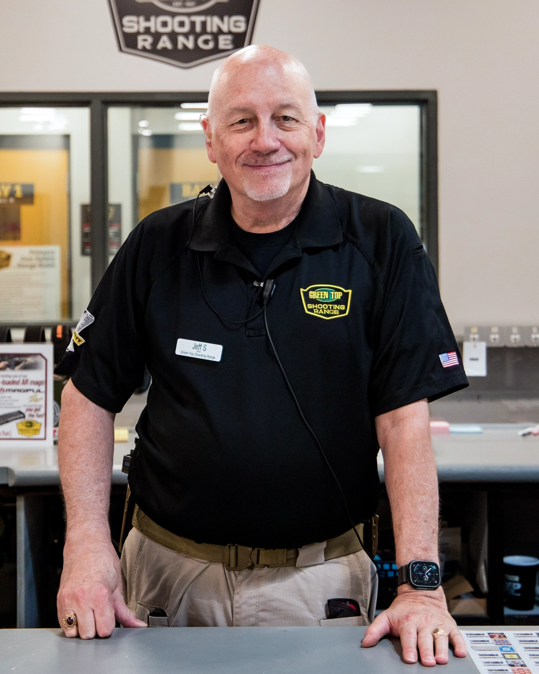 Jeff Summers - Retail Sales Supervisor, wearing a black Green Top Shooting Range shirt, stands behind a counter, leaning forward with one hand resting on the surface in a shooting range facility.