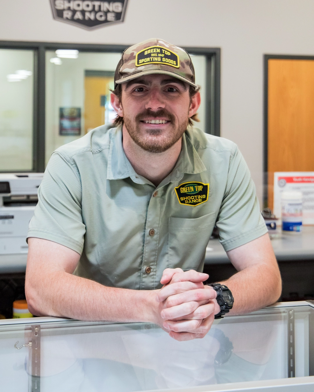 Kevin Dunne - Training and Membership Manager, wearing a Green Top Sporting Goods cap and shirt, stands behind a counter smiling with hands clasped. Office doors and shooting range signs are visible in the background.