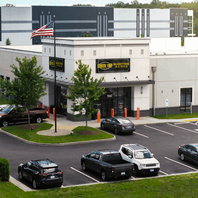 A modern, single-story commercial building with a green top shooting range exterior and a U.S. flag in front, surrounded by parked cars and trees, with a large warehouse or office building visible in the background.